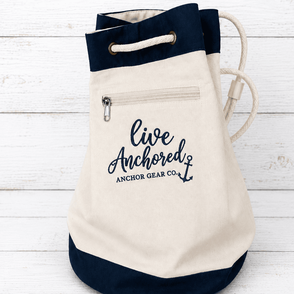 Canvas drawstring bag with navy accents and 'Live Anchored' text on a white wooden background