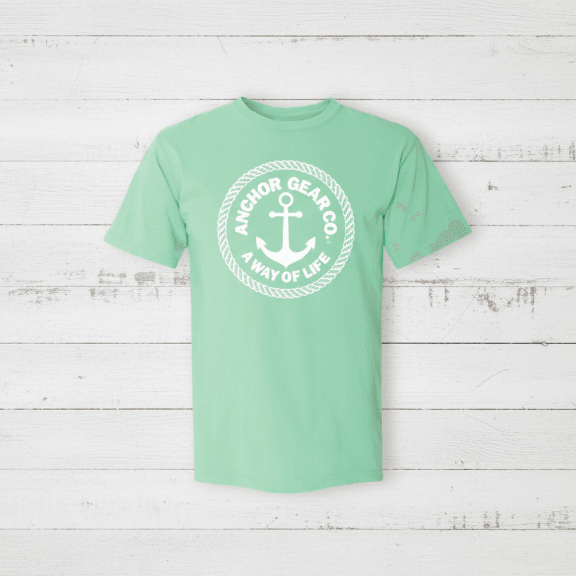 A green crew neck t-shirt with the Anchor Gear Co. logo printed in the center, displayed on a wooden jetty with trees and water in the background.