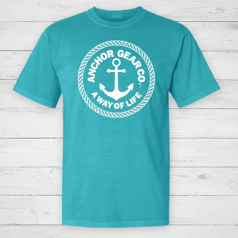 A green crew neck t-shirt with the Anchor Gear Co. logo printed in the center, displayed on a wooden jetty with trees and water in the background.