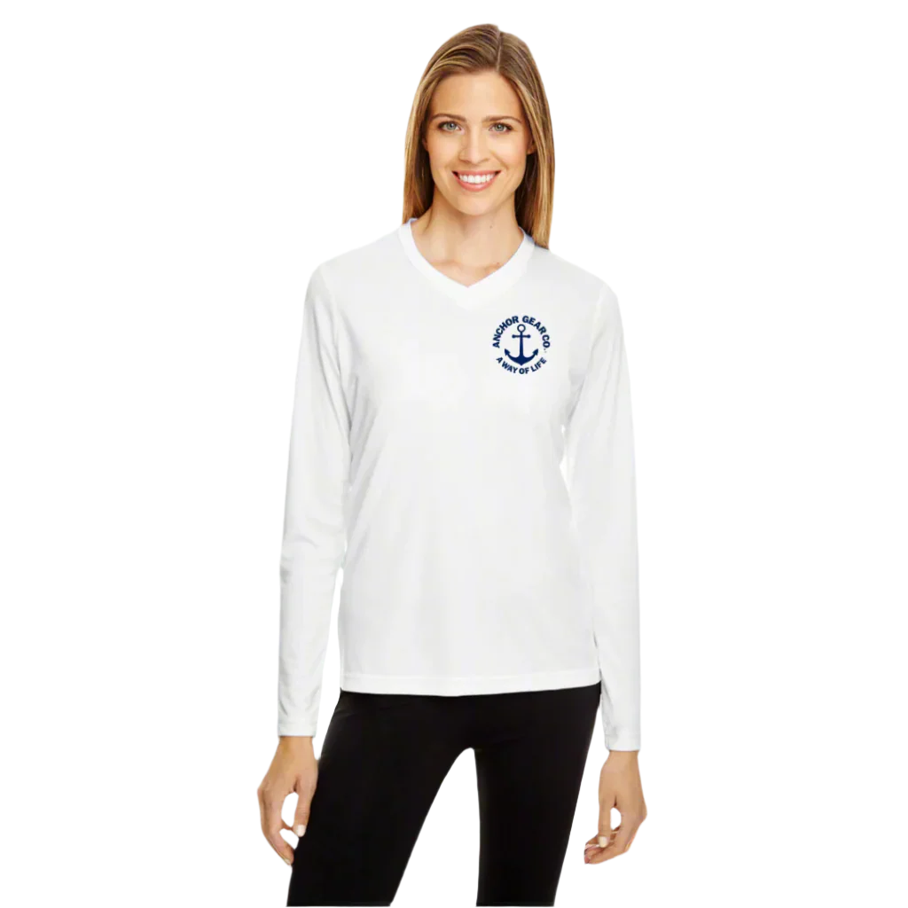 A woman wearing a white long sleeve UV shirt with the Anchor Gear Co. logo on the front left chest, standing near a marina with boats in the background.