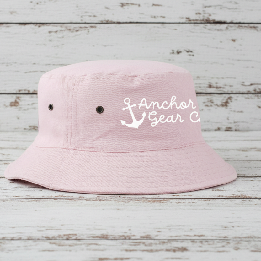 A pink Youth Nautical Bucket Hat with a whimsical Anchor Gear Co. logo on the front, displayed on a wooden surface.