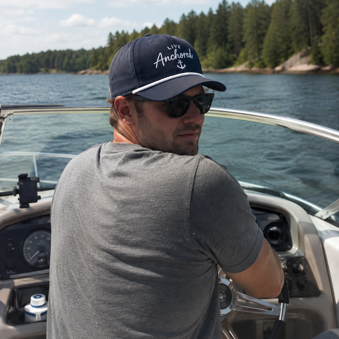 A man wearing a navy blue 'Anchors Avo' baseball cap and sunglasses, driving a boat on a lake with a forested shoreline in the background.